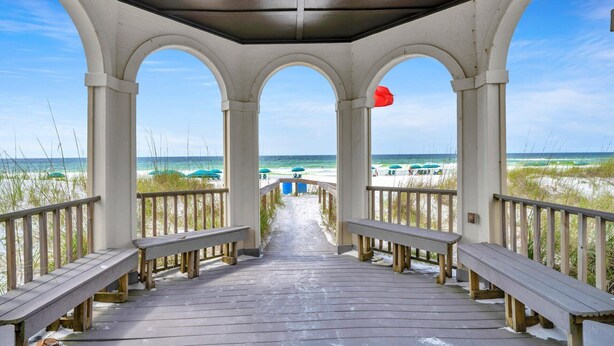 Une terrasse en bois couverte, dotée de bancs, qui mène à une plage ombragée de parasols.