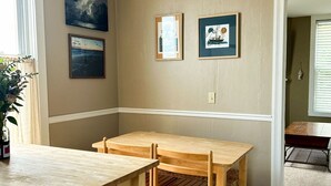 Une pièce avec une table et des chaises en bois, une fenêtre avec des rideaux et des œuvres d’art accrochées au mur.