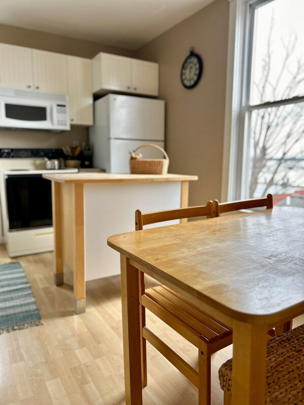 A kitchen with wooden cabinets, a microwave, and a refrigerator. There is a dining table with chairs and a basket on the counter. - Great Water Inn (Bayfield)