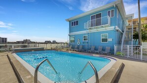 A clear blue swimming pool with a metal ladder, surrounded by lounge chairs and a two-story building with balconies.