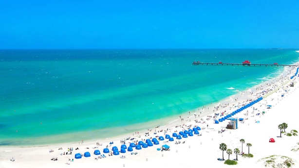A beach with a pier extending into the ocean, numerous umbrellas, and people enjoying the beach.