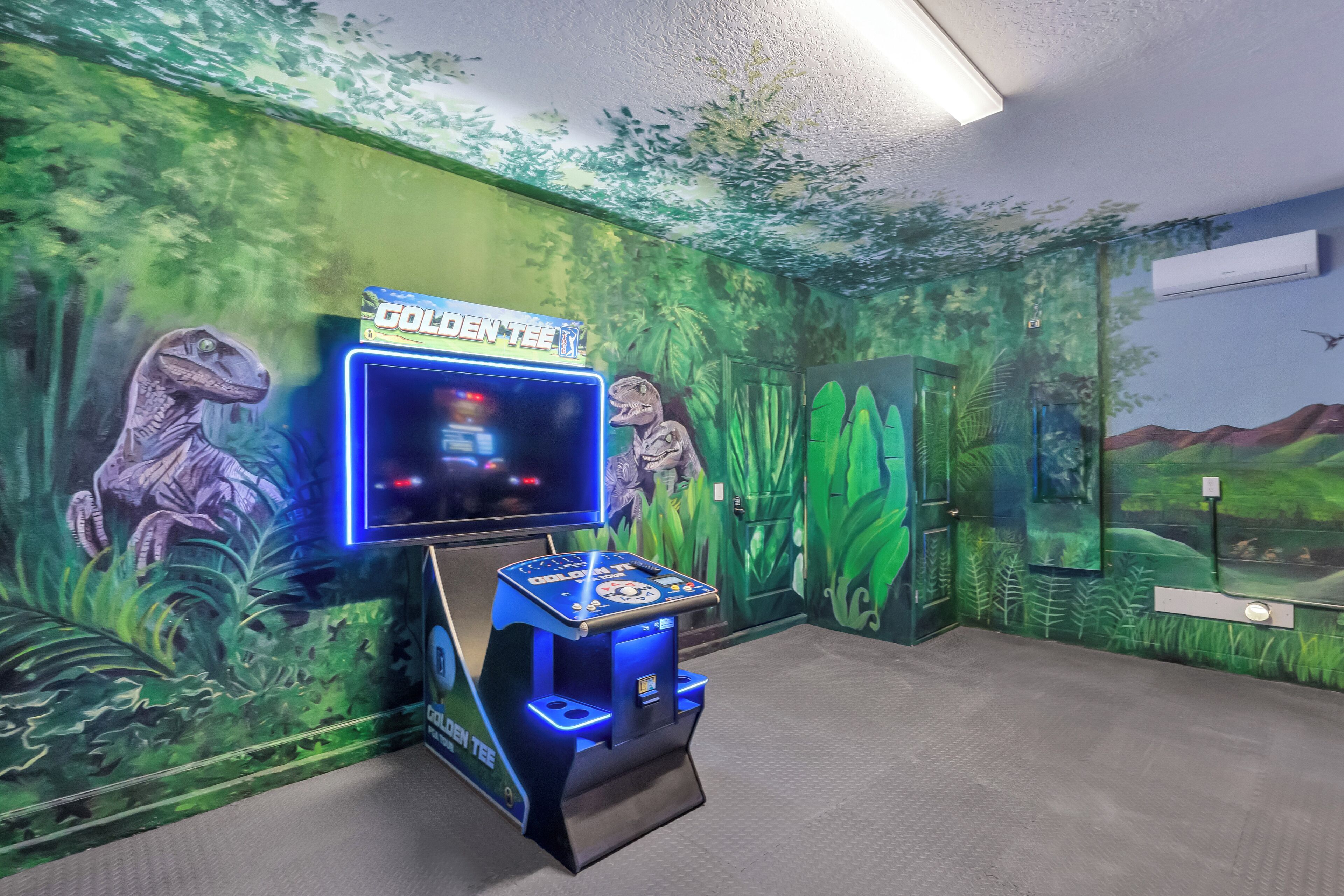 Ein Innenraum mit Wandgemälde zum Thema Dinosaurier, einem 'Golden Tee' Arcade-Automaten und einem großen Bildschirm.