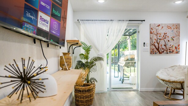 A modern living room with a flat-screen TV displaying various streaming services, a large potted plant, a white chair, and a view of the outdoors through a glass door. - Modern Relaxing Retreat w/ Pool, Fire pit, Game room (Orlando)