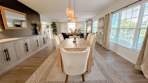 A spacious dining area with a wooden table and white chairs, a large window with blinds, and a hanging light fixture. - Modern Relaxing Retreat w/ Pool, Fire pit, Game room (Orlando)