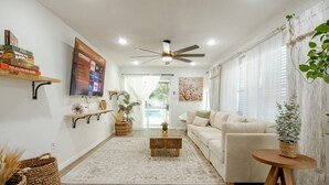 A modern living room with a white sofa, a coffee table, and a TV mounted on the wall. There are shelves with books and decorative items, and a ceiling fan. - Modern Relaxing Retreat w/ Pool, Fire pit, Game room (Orlando)