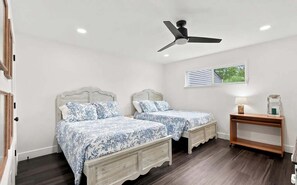 Quarto com duas camas, um ventilador de teto, mesa de cabeceira com abajur e janela com persianas.