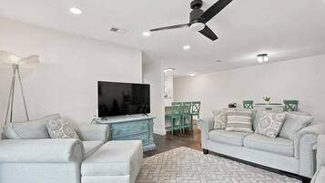 A modern living room with a flat-screen TV, two sofas, a ceiling fan, and a dining area with chairs.