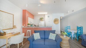 A modern living room with a blue sofa, white chairs, a kitchen area with a pink accent wall, and a dining table with blue chairs.