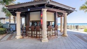 A covered outdoor seating area with wooden chairs and tables, a closed shutter, and a view of the beach.