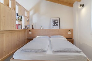 A wooden bed with white linens, a wooden bookshelf, a framed picture on the wall, and a nightstand with a lamp.