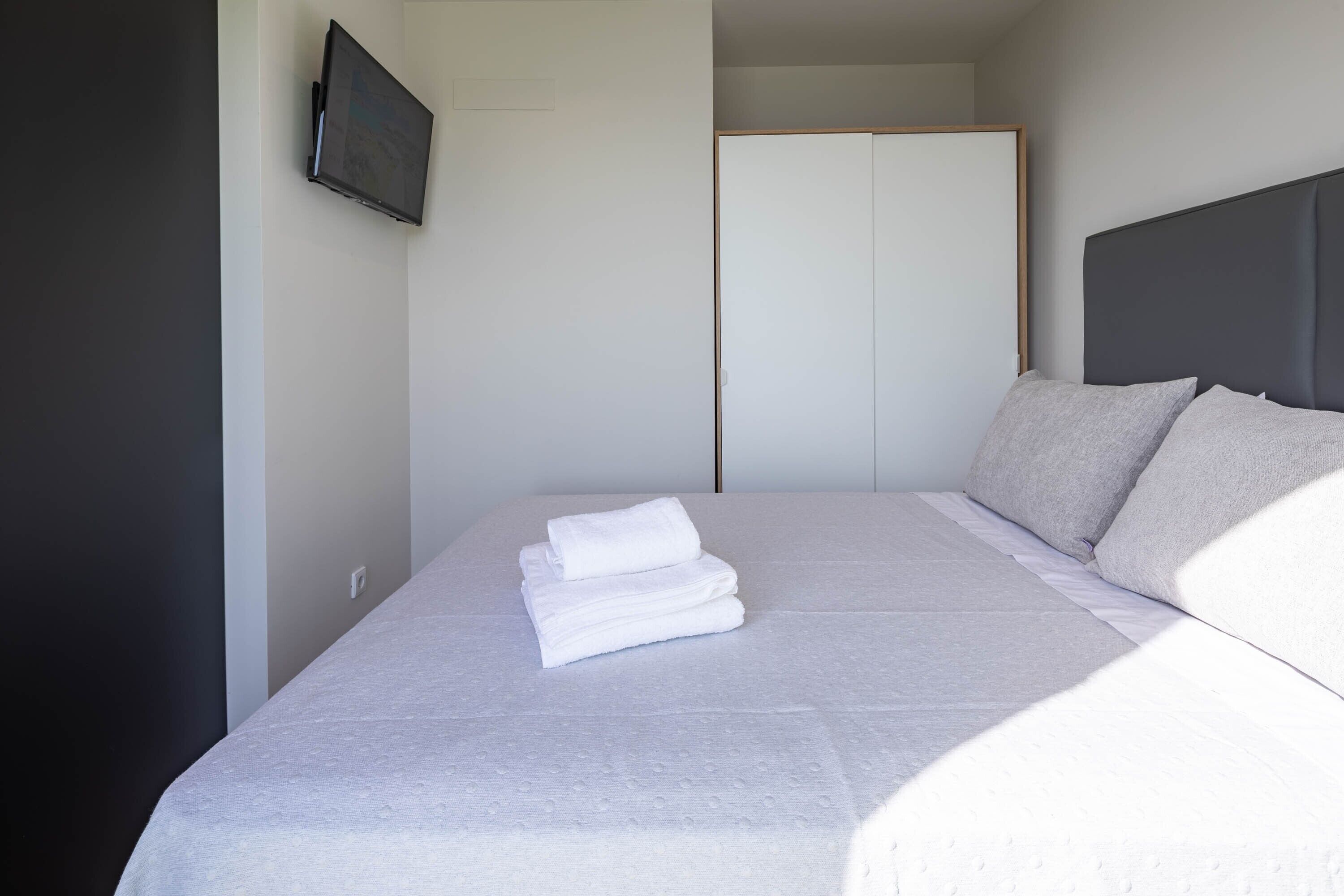 A neatly made bed with folded white linens, a grey headboard, and a white wardrobe in the background.