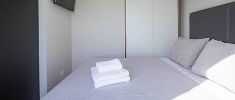 A neatly made bed with folded white linens, a grey headboard, and a white wardrobe in the background.