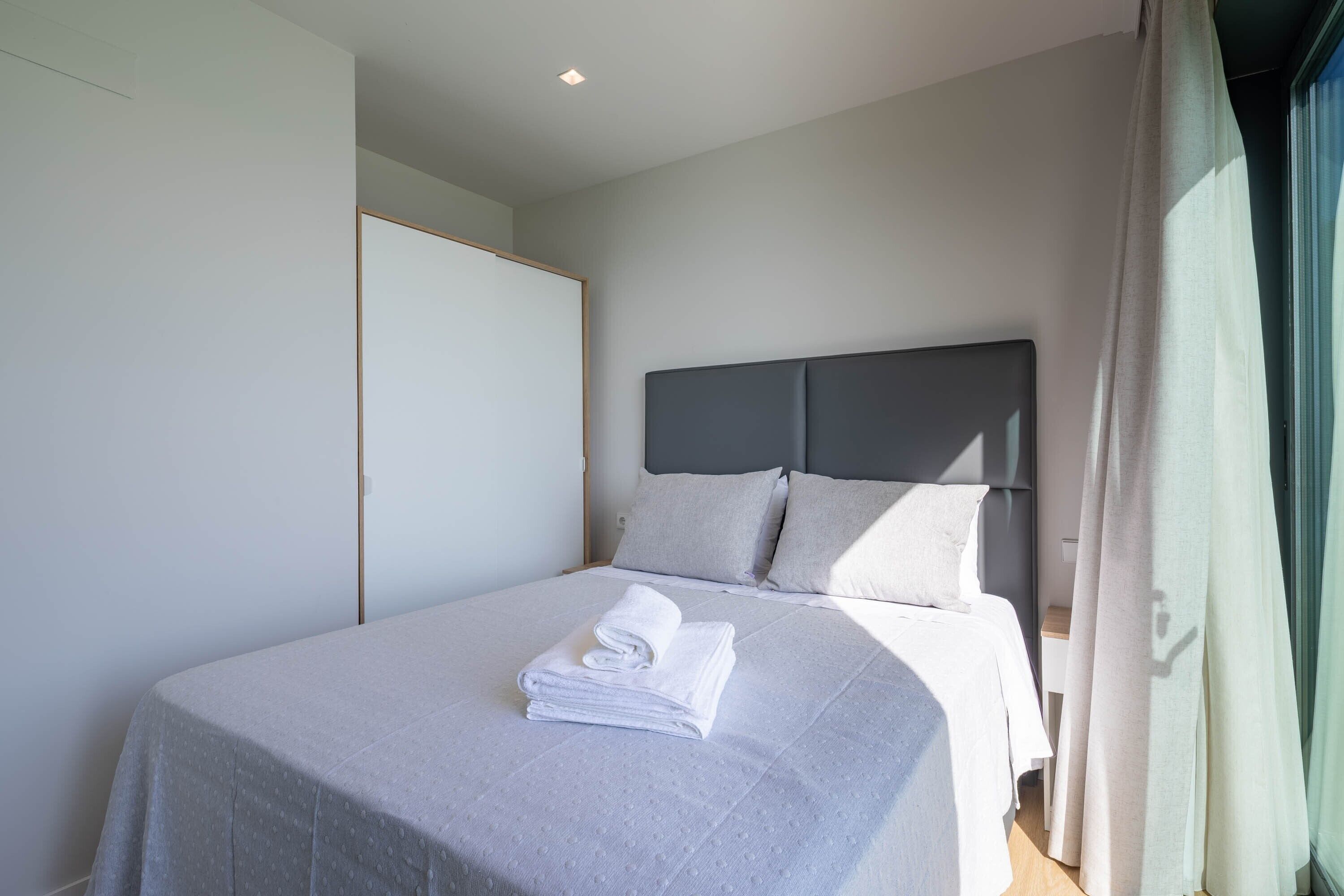 A neatly made bed with a folded towel, a grey headboard, and a window with curtains.