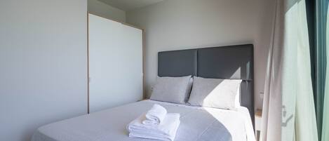 A neatly made bed with a folded towel, a grey headboard, and a window with curtains.