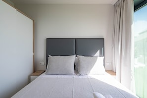 Una cama con cabecera gris, dos almohadas y una colcha blanca en una habitación con ventana y cortinas.