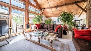 A spacious living area with a wooden ceiling, large windows, a sofa, a coffee table, and potted plants.