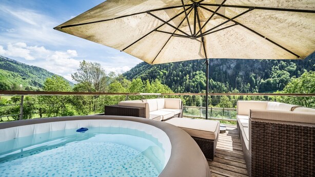 A hot tub on a deck with outdoor furniture and a view of a forested mountain.