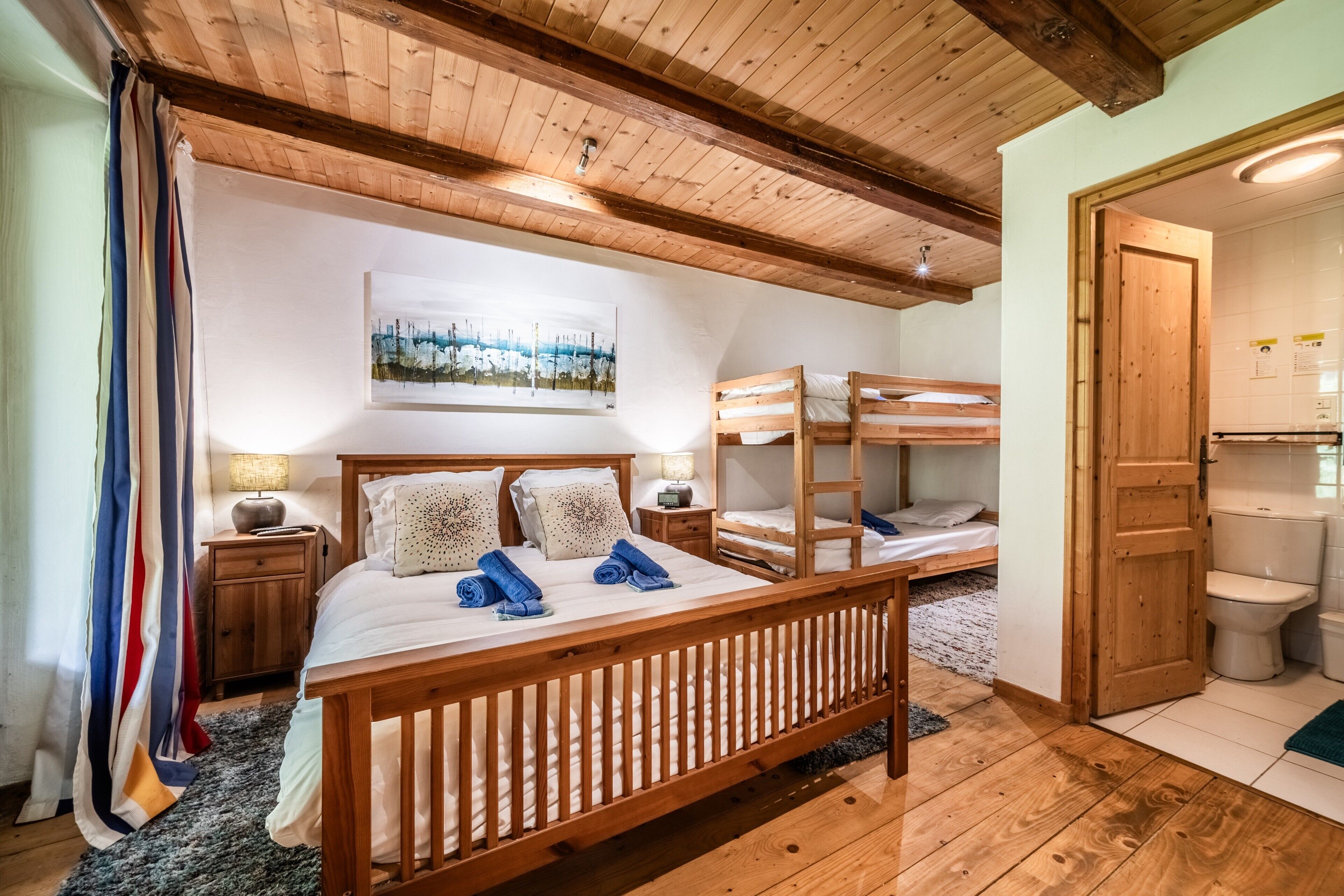 Une chambre à coucher avec un lit en bois, des lits superposés, une table de chevet, une lampe, un tableau, et une salle de bain avec des toilettes et un lavabo.