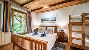 Habitación de madera con una cama, literas, una ventana con cortinas, un cuadro y una mesita de noche con lámpara.