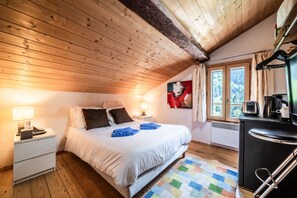 Ein Schlafzimmer mit Holzboden, einem Bett mit weißer Bettwäsche, einem Nachttisch mit Lampe, einem Fenster mit Vorhängen und einer Küchenzeile.