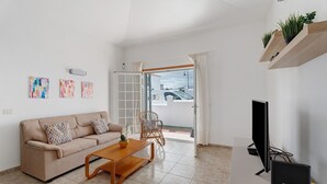 Ett vardagsrum med en soffa, ett soffbord och en tv. Det finns växter på en hylla, konstverk på väggen och en utsikt genom en glasdörr.