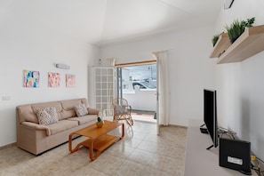 Un salon avec un canapé, une table basse et un téléviseur. On y trouve des plantes sur une étagère, des œuvres d’art accrochées au mur et une vue à travers une porte vitrée.