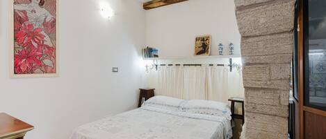 Un dormitorio con cama, cabecero de madera, pared de piedra, ventana con mosquitero y un cuadro en la pared.