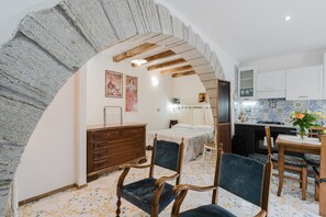 Una habitación con cama, cómoda de madera, sillas, una mesa y una zona de cocina con armarios y salpicadero de azulejos.