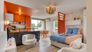 A modern hotel room with a kitchenette, a bed with striped bedding, a white armchair, and a dining area with orange chairs. - Apartment "Sunny Day" with Private Terrace and Air Conditioning (La Grande-Motte)