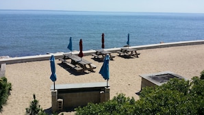 Une plage avec des tables de pique-nique en bois et des parasols bleus, une promenade en béton et une vue sur la mer.
