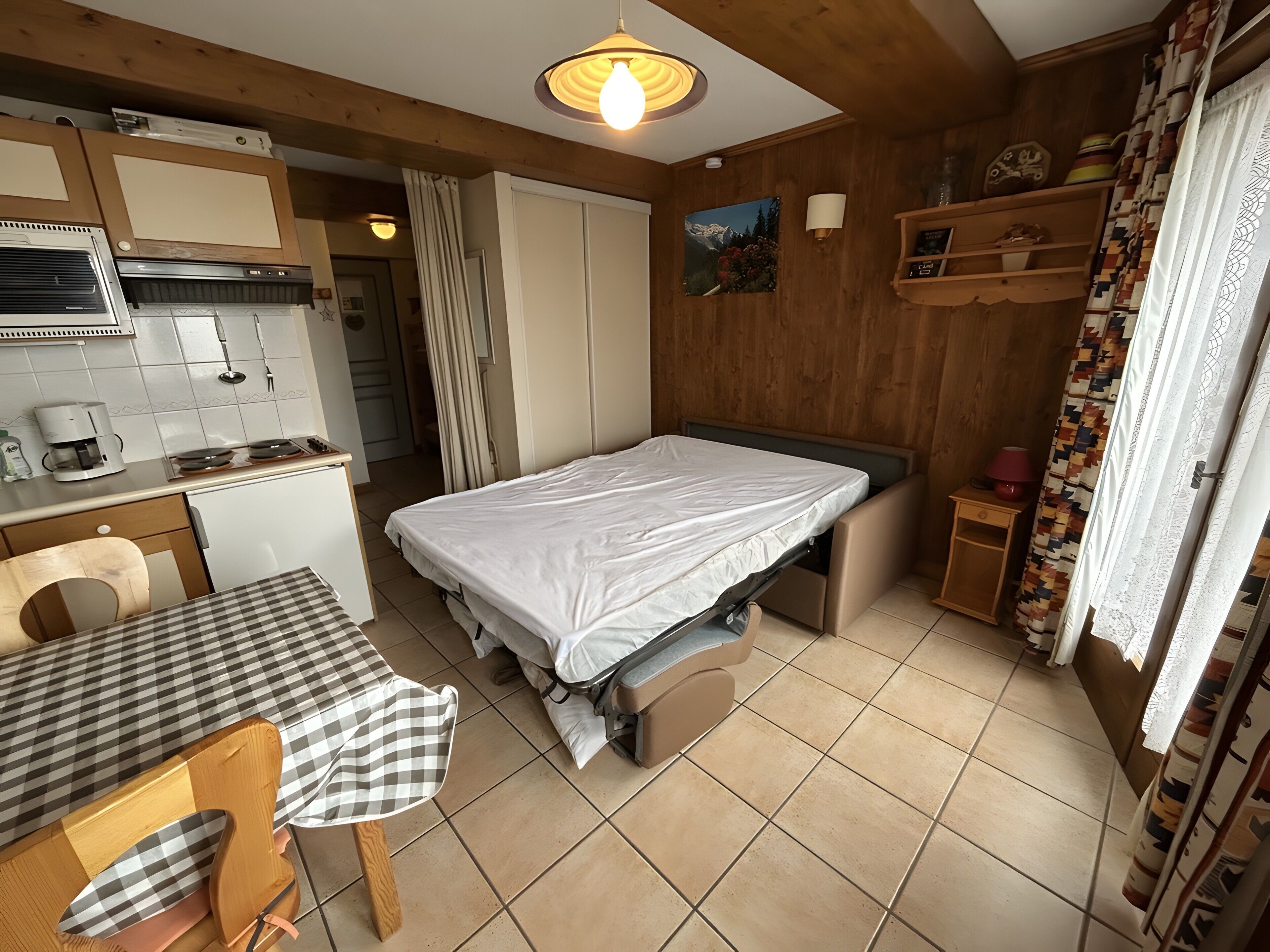 A compact room with a bed, a small table, a chair, a kitchenette, and a ceiling light.