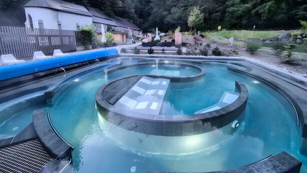 A circular hot spring pool with a surrounding deck, set in a forested area with a house in the background.