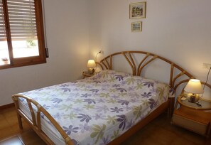 Un lit simple avec une couvre-lit à motifs floraux, deux lampes de chevet, une tête de lit en bois et une fenêtre avec des stores.