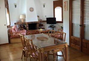 Un coin repas avec une table en verre et des chaises en osier, une cheminée, un téléviseur et une porte avec des stores.