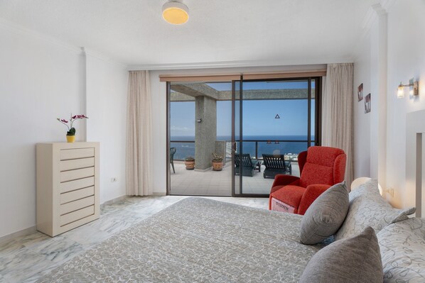 A bedroom with a bed, a red chair, a dresser, and a balcony with a view of the sea.