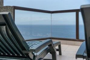 A balcony with a view of the ocean, a lounge chair, and a table.