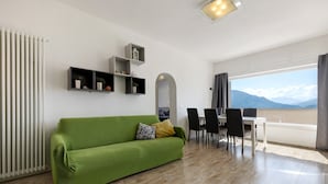 A modern living room with a green sofa, a dining area with chairs, a white table, and a view of mountains through a large window. - Villa 'Renè' with Private Terrace, Wi-Fi and Air Conditioning (Tenno)