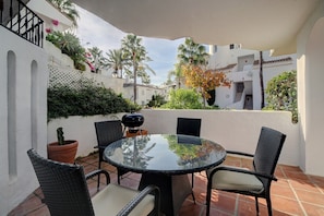Ett runt glasbord med fyra flätade stolar, en grill och krukväxter på en terrass med utsikt över de omkringliggande byggnaderna.