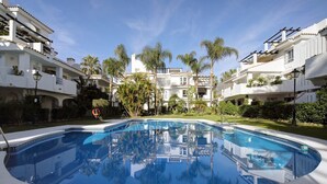 A swimming pool surrounded by white buildings with balconies, palm trees, and a clear sky.