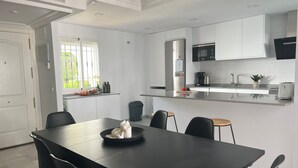 A modern kitchen with white cabinets, a stainless steel refrigerator, a microwave, and a coffee maker. There is a dining table with chairs and a window with a view of greenery outside.