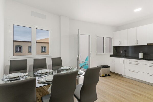 Una cocina moderna con armarios blancos, encimera negra y una mesa de vidrio para cuatro comensales.