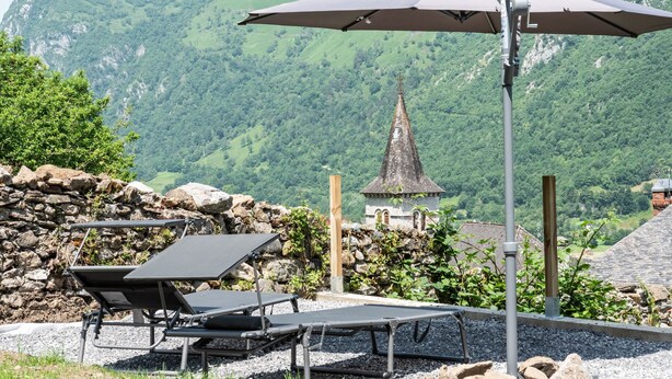 Eine Terrasse mit Sitzgelegenheit, Blick auf einen Berg und ein kleines Gebäude mit spitzer Dachform.