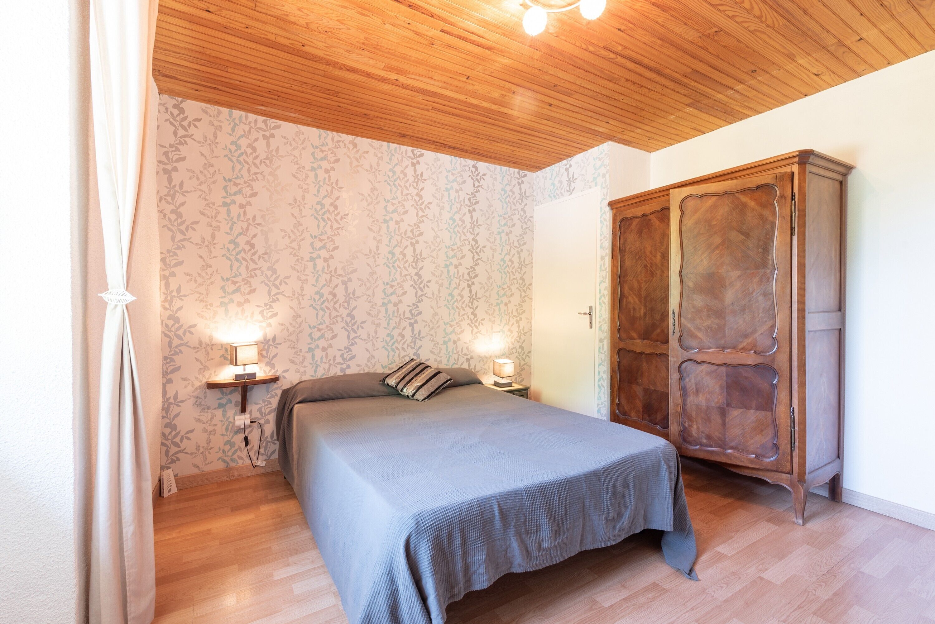 A bedroom with a bed, bedside lamps, a wooden wardrobe, and patterned wallpaper.
