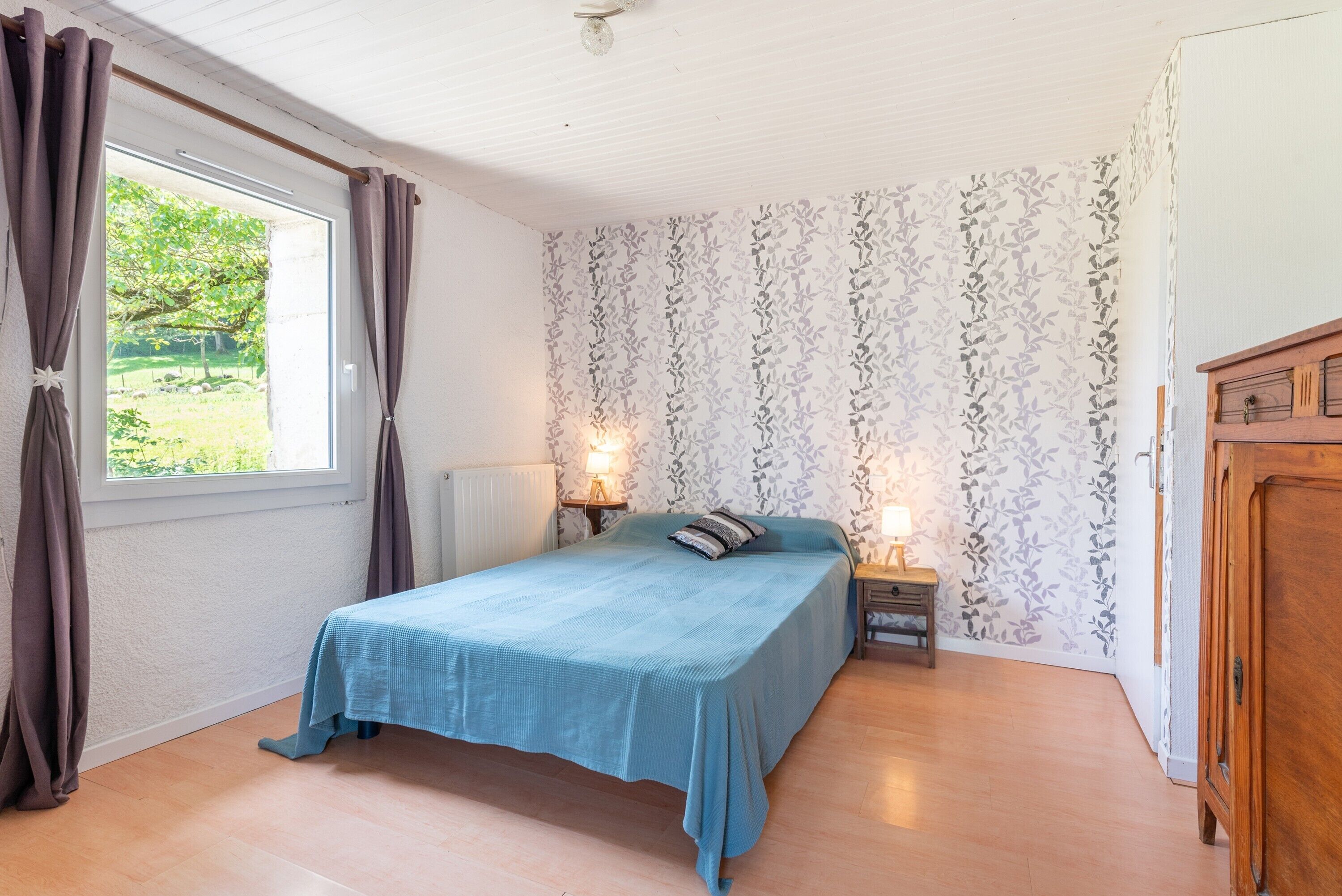 A bedroom with a bed, curtains, a nightstand, a lamp, a radiator, and a view of greenery outside.