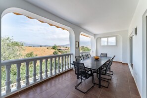 A balcony with a dining table and chairs, a white railing, and a view of a landscape with mountains and trees.