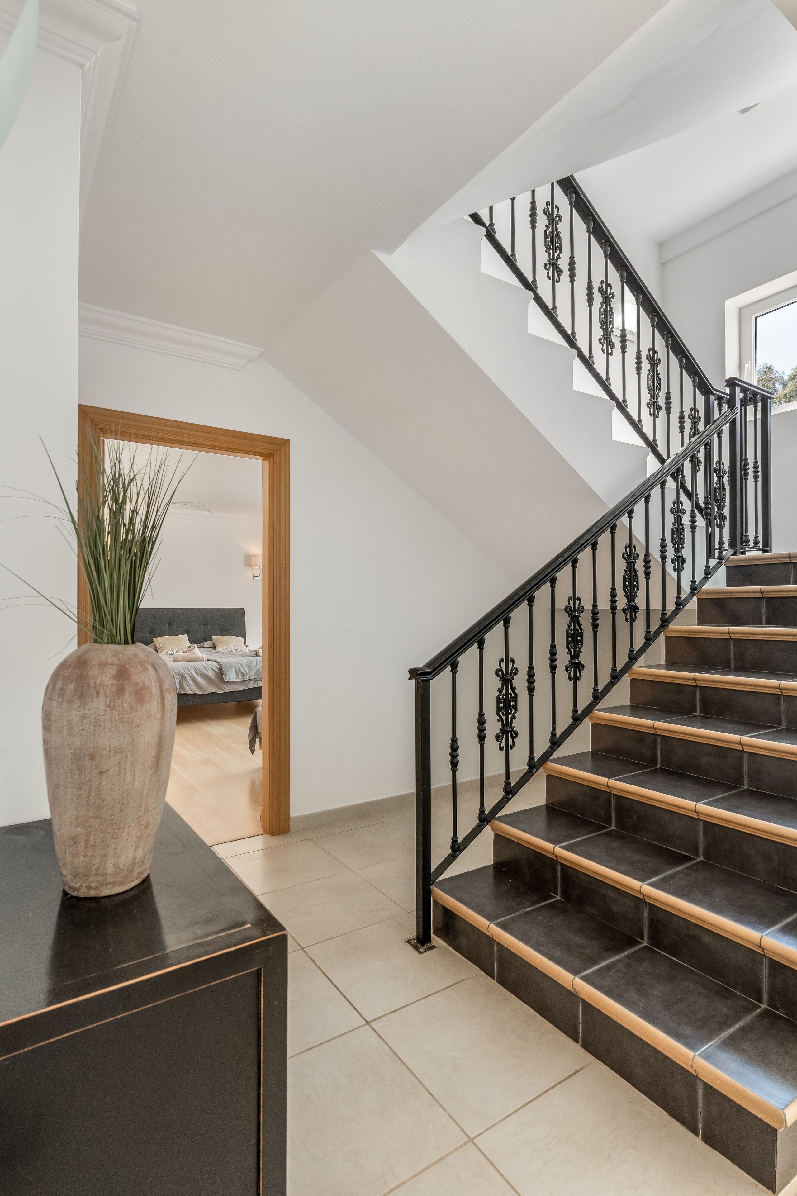 A staircase with black balusters and beige steps, leading to a bedroom with a bed and a plant.