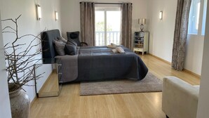 Un dormitorio con cama, mesitas de noche, una silla, lámpara, ventana con cortinas y vista al exterior.