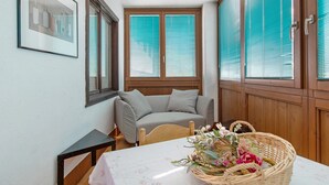 Ein Zimmer mit einer Couch, einem Tisch mit Blumenmuster-Tischdecke, einem Korbwägelchen mit Blumen und einem gerahmten Bild an der Wand.