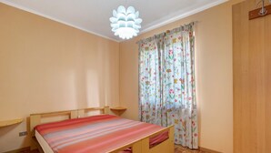 Un dormitorio con cama, cabecera de madera, una mesita de noche pequeña, una lámpara colgante y una ventana con cortinas estampadas.