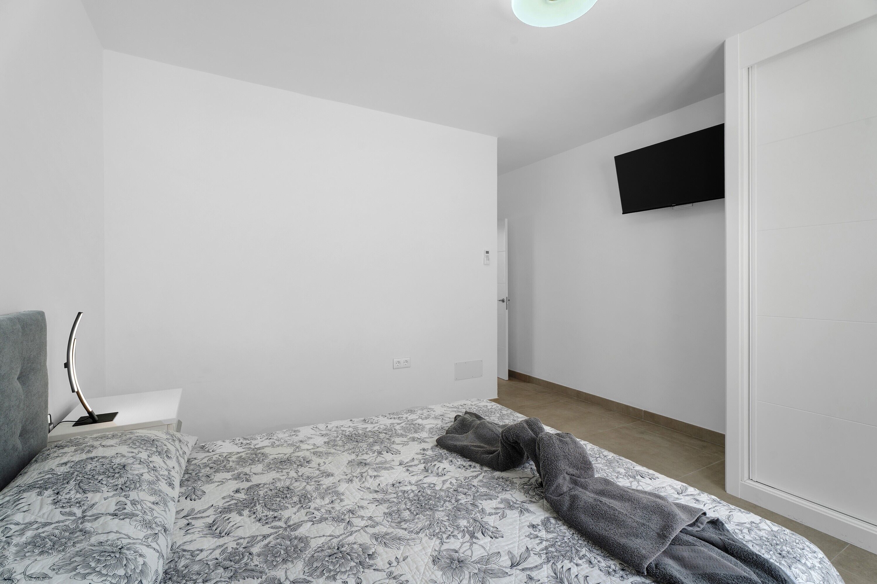 Une chambre à coucher avec un lit, un téléviseur à écran plat fixé au mur et une table de chevet avec une lampe.